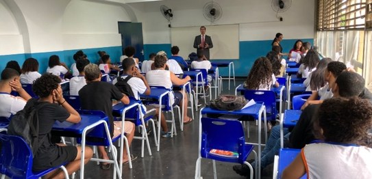 Descrição da imagem: Em pé, o juiz veste um terno e está de frente para os(as) alunos(as). Os(as...