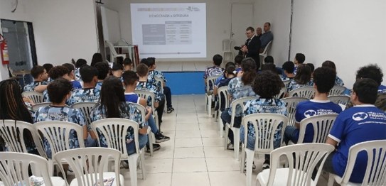 Descrição da imagem: O juiz veste um terno preto e segura um microfone em cima do palco. As(os) ...